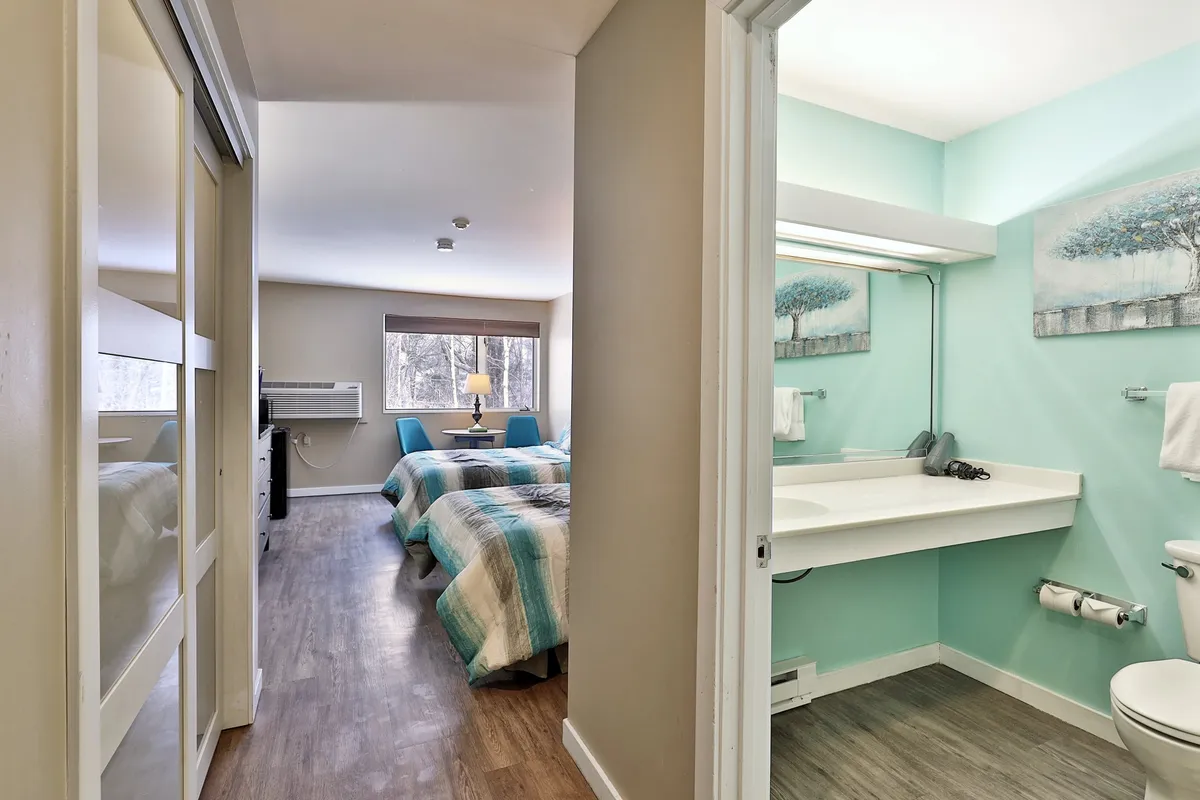 This image displays a welcoming interior with two beds featuring striped comforters, a visible bathroom sink area, and decorative wall art. The soft lighting and inviting arrangement evoke a restful and clean atmosphere. The sink area subtly references personal grooming and comfort, while the beds suggest restful nights.