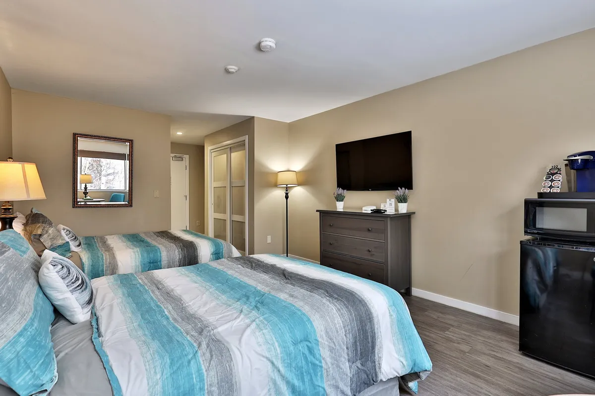 The image highlights a comfortable area with two beds dressed in striped bedding, a wall-mounted television, a nightstand lamp, and a mini-fridge. The scene feels inviting and well-equipped for both relaxation and productivity.