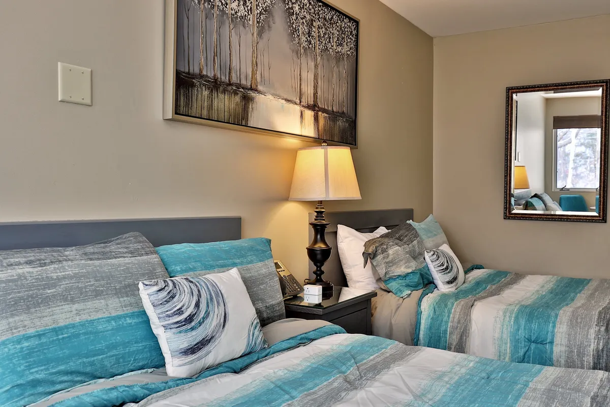 The image showcases a tranquil interior of a renovated studio with two inviting beds adorned in blue and gray striped bedding, softly illuminated by a bedside lamp. Above the beds hangs a large, calming painting, and a mirror on the wall adds to the room’s sense of warmth and space. The atmosphere suggests comfort, relaxation, and a welcoming retreat after a day on the slopes.