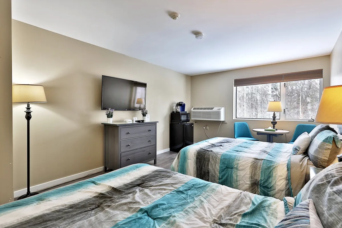 A cozy studio interior with two beds adorned in colorful bedding, a wall-mounted television, a softly glowing lamp, and a small table set by the window. The space looks inviting for relaxing, watching TV, or enjoying a quiet moment with coffee.