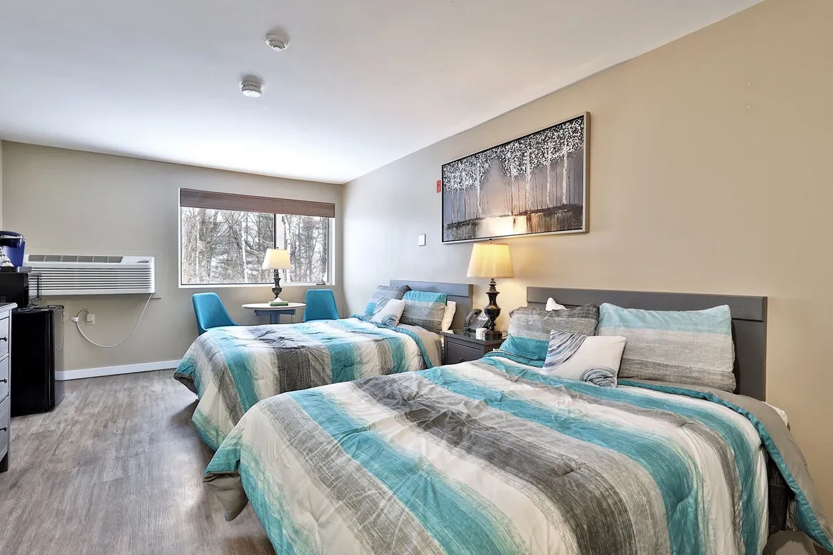 This image highlights two beds with striped bedding, a bedside lamp, and a painting on the wall. The scene exudes a tranquil, cozy ambiance, with soft natural light filtering in. The arrangement offers a clear view of the sleeping area, showcasing the comfort and warmth of the studio layout.