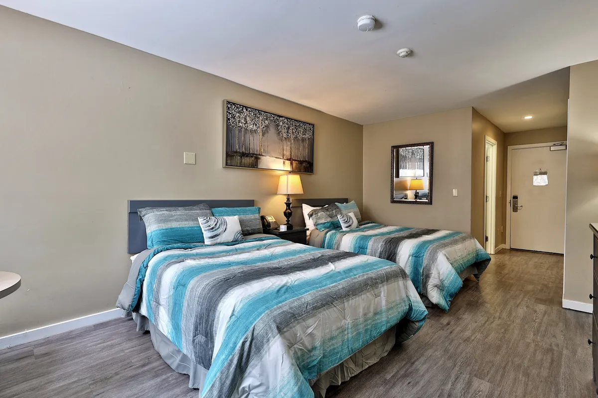 A comfortable hotel studio with two beds featuring aqua and gray bedding, a softly lit nightstand, and framed artwork. The inviting layout encourages relaxing conversations or planning the next day’s adventures, capturing the in-between moments of a lively getaway.