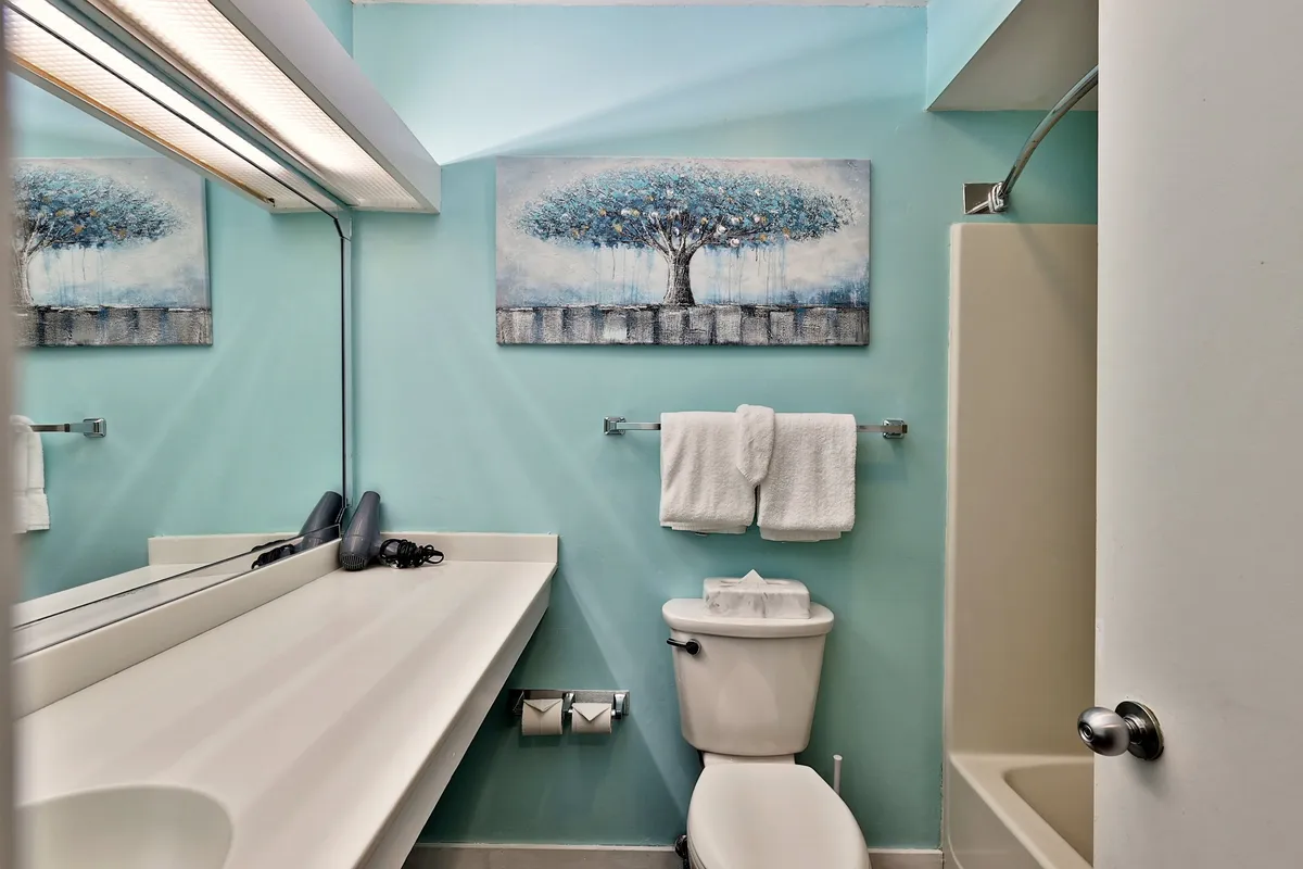 This image features a large mirror, a tree-themed painting, crisp white towels, and the clean lines of a well-lit bathroom sink. The serene mood is complemented by subtle decorative touches, giving the bathroom a boutique hotel feel.