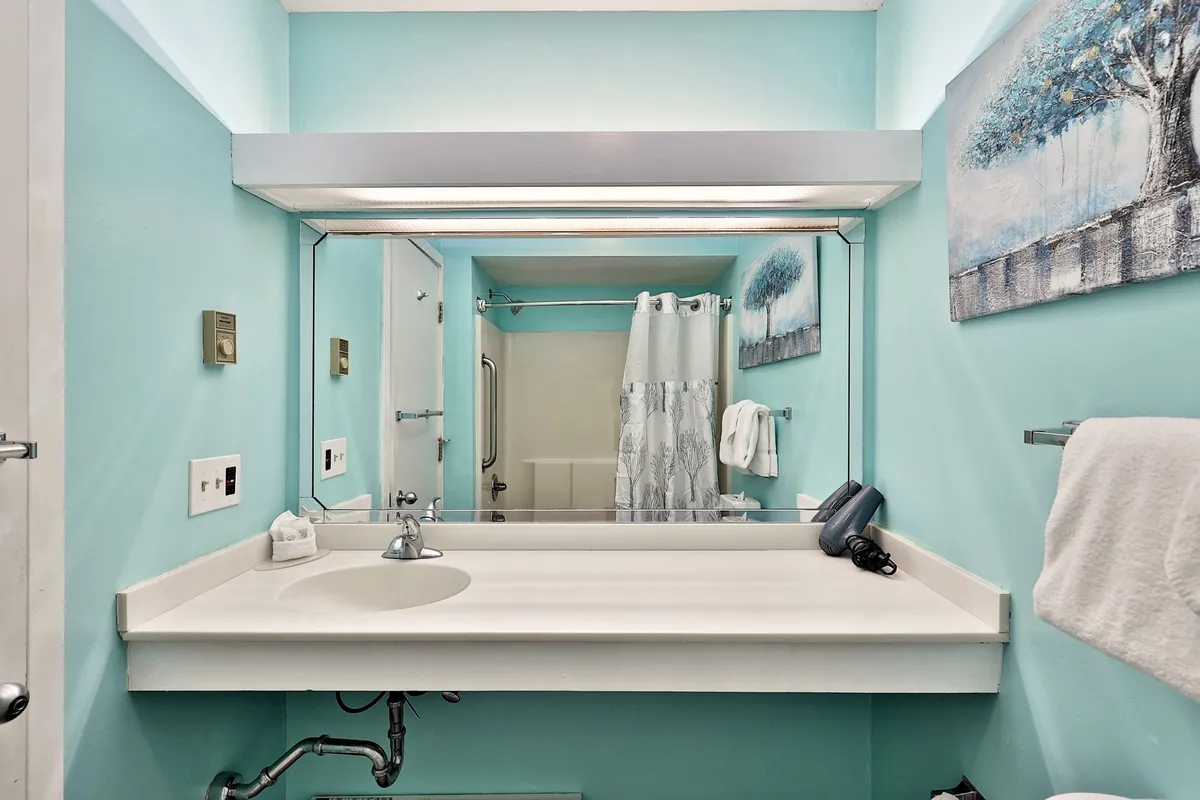 A calm, sunlit bathroom featuring a mirror above a sink, soft towels neatly arranged, subtle wall art for a personal touch, and a shower curtain. Morning sunlight gently fills the space, creating a tranquil atmosphere ideal for unwinding after outdoor activities.