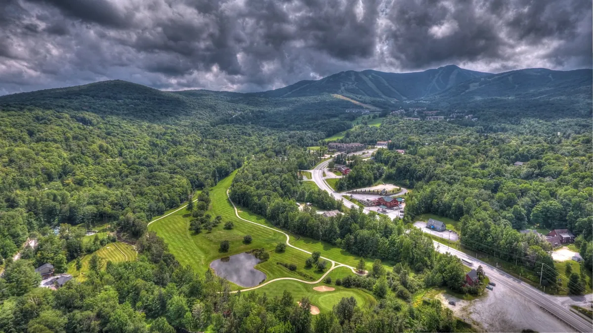 A sweeping panoramic view featuring rolling mountains, a golf course, a tranquil pond, and local roadways, capturing the broader Killington outdoor scene and the area's year-round recreational appeal.